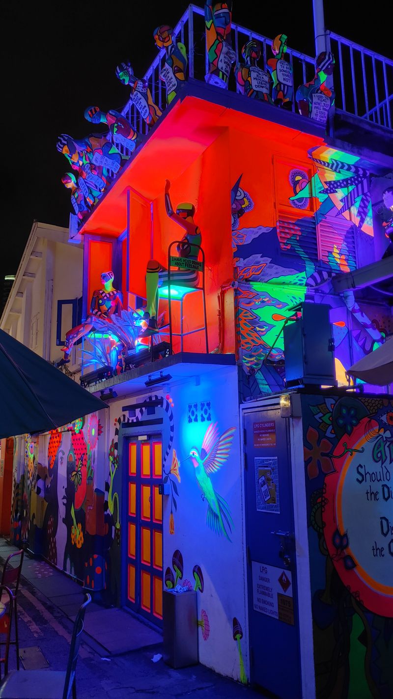 a bar building with blue and red lights on the outside with paintings of birds and manequins sitting in chairs waving, along with other small figures hanging from the top floor balcony railings