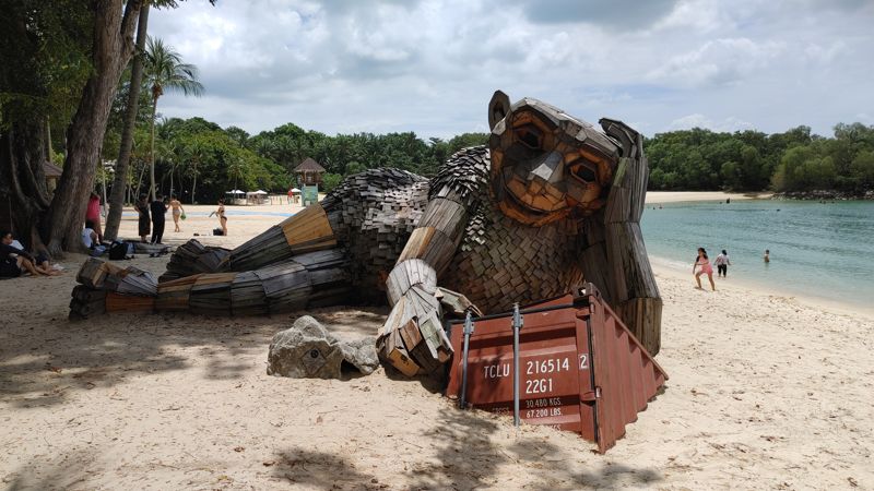 art sculpture of a giant monkey-like animal made of wood panelings lying on its side and looking at the corner of a shipping container buried in the sand
